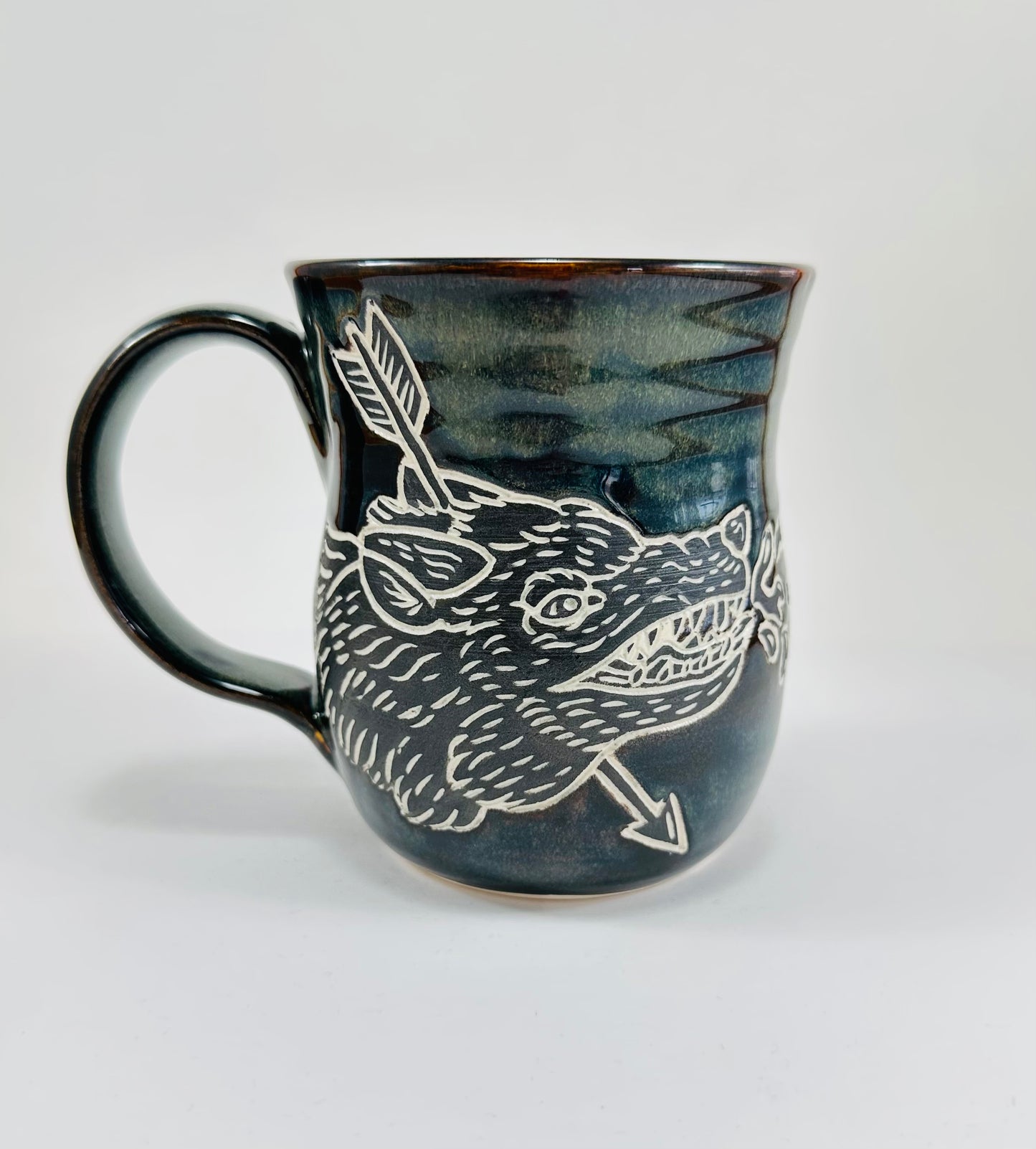 A handmade ceramic mug with an illustration of a wolf head and arrow on it. The mug is glazed in a beautiful dark blue/green glaze an has a smooth comfortable handle. The mug is made from porcelain clay.