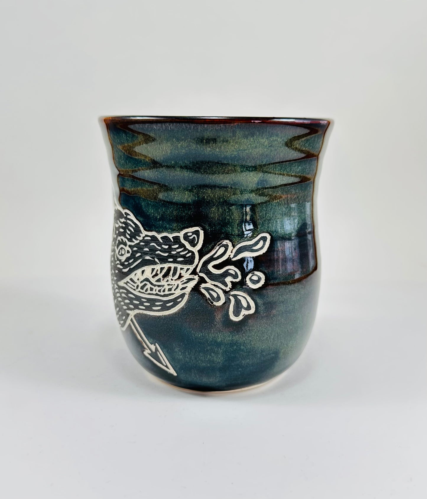 A handmade ceramic mug with an illustration of a wolf head and arrow on it. The mug is glazed in a beautiful dark blue/green glaze an has a smooth comfortable handle. The mug is made from porcelain clay.