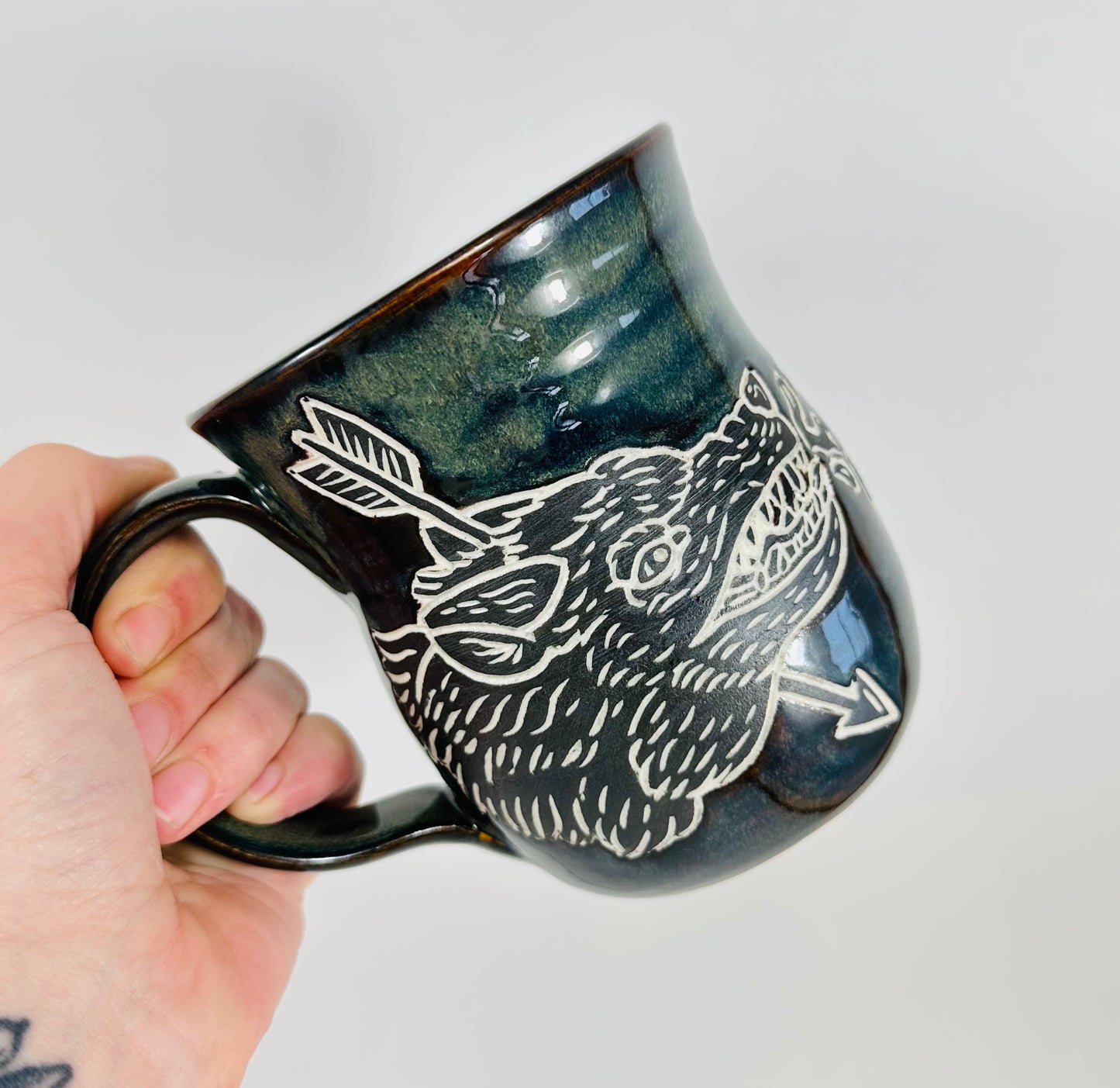 A handmade ceramic mug with an illustration of a wolf head and arrow on it. The mug is glazed in a beautiful dark blue/green glaze an has a smooth comfortable handle. The mug is made from porcelain clay.