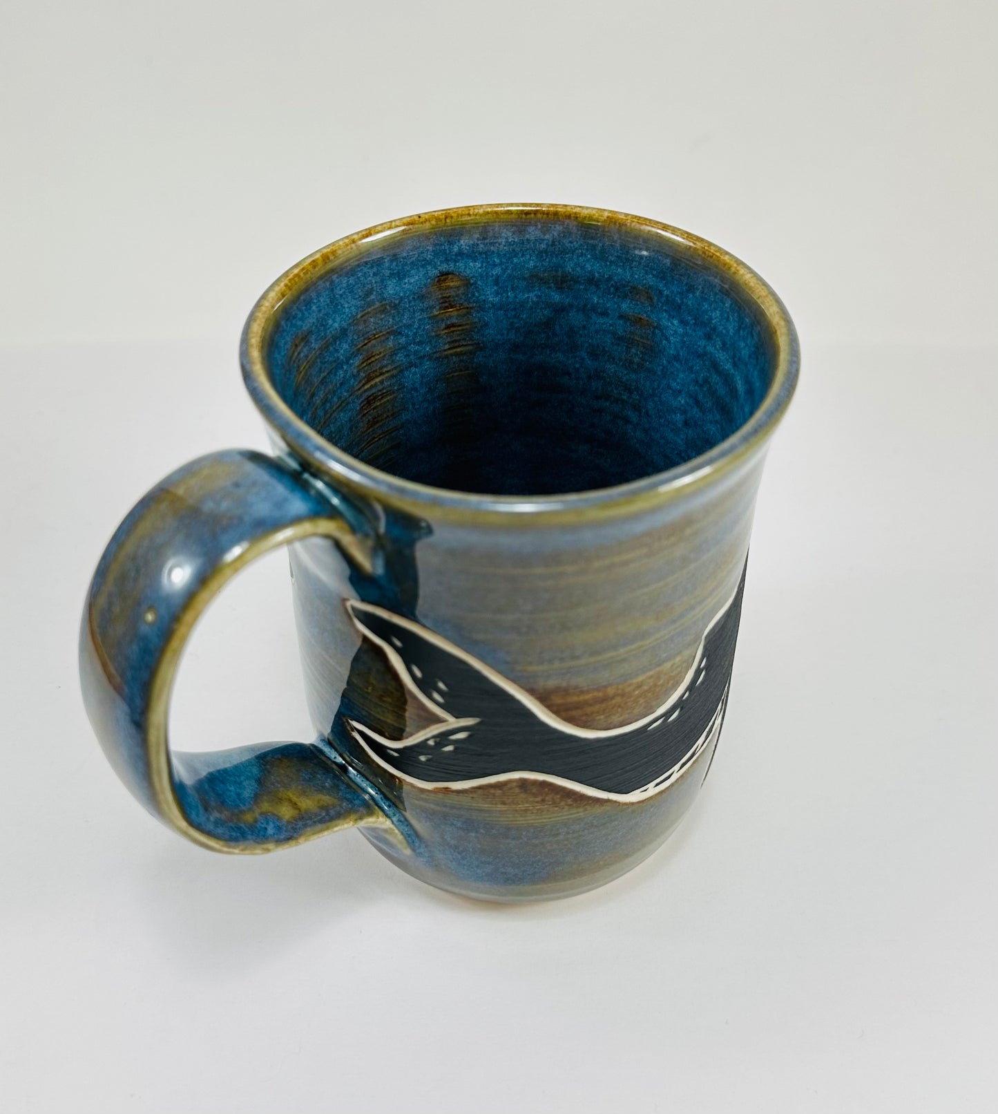 A handmade ceramic mug with a blue whale illustration on it and fish. The mug is made from porcelain clay and has a beautiful blue and brown glaze.