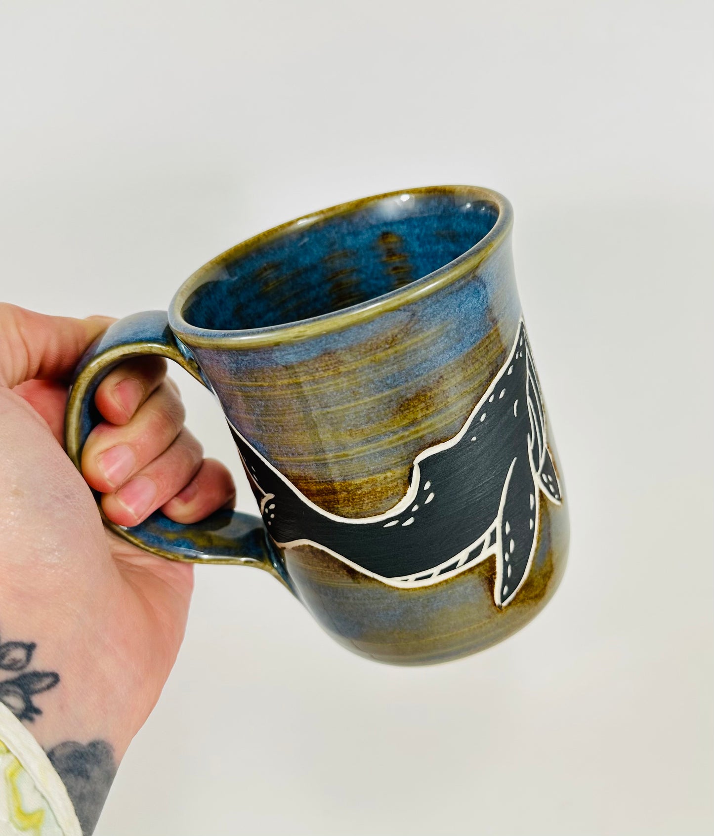 A handmade ceramic mug with a blue whale illustration on it and fish. The mug is made from porcelain clay and has a beautiful blue and brown glaze.