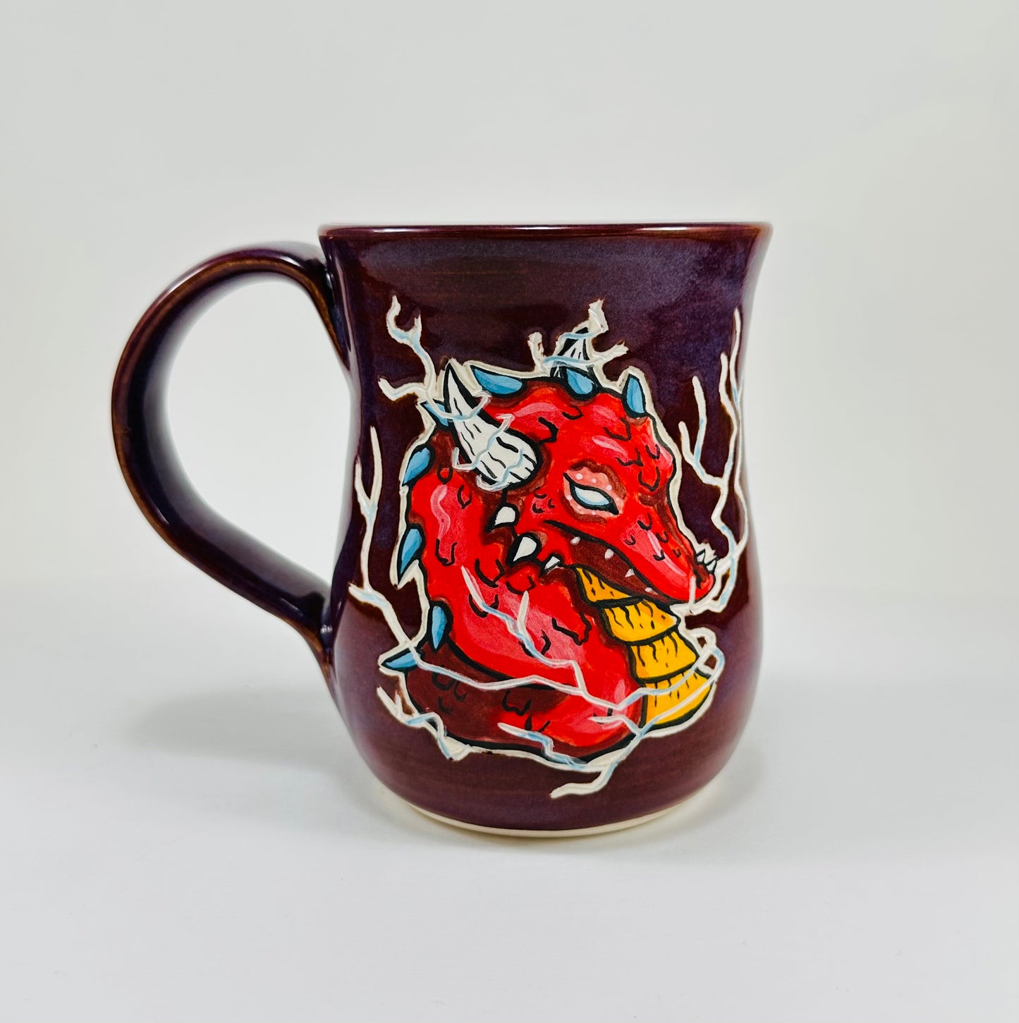 A handmade ceramic mug with a red dragon illustration on it. The mug is glazed in a beautiful purple glaze and is made from porcelain clay.