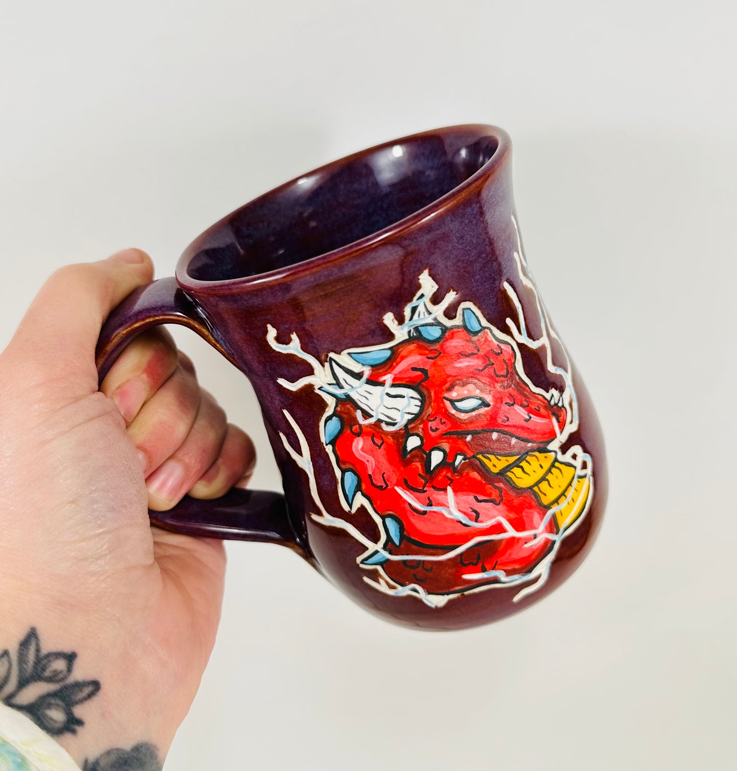 A handmade ceramic mug with a red dragon illustration on it. The mug is glazed in a beautiful purple glaze and is made from porcelain clay.