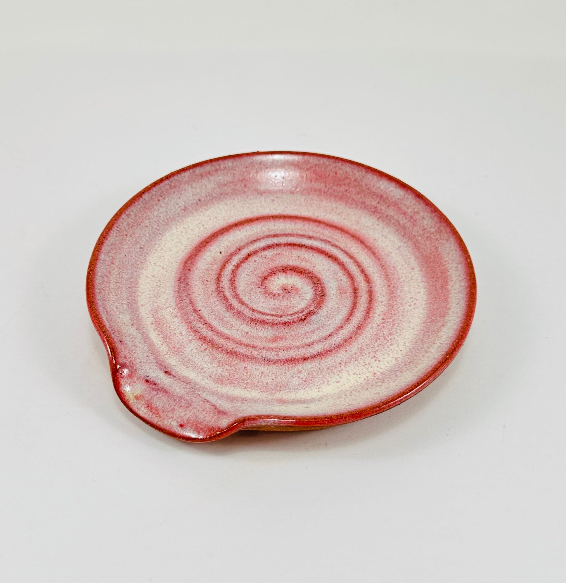 A handmade ceramic spoon rest in a beautiful pink glaze on red clay.