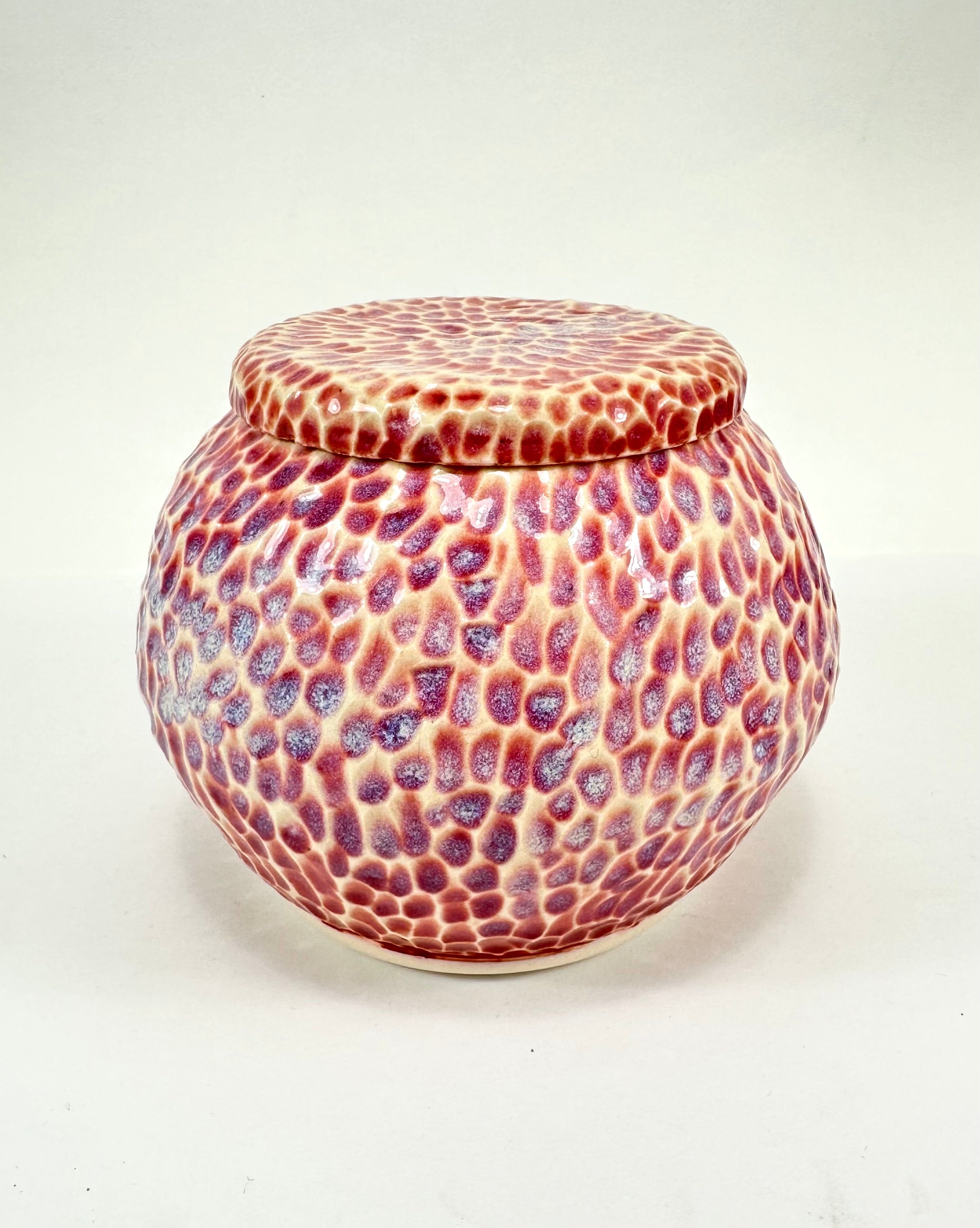 A handmade ceramic sugar jar, The sugar jar is glazed in a beautiful pink, blue, beige and red glaze. It is carved all over to add texture. The sugar jar is round with a flat lid.
