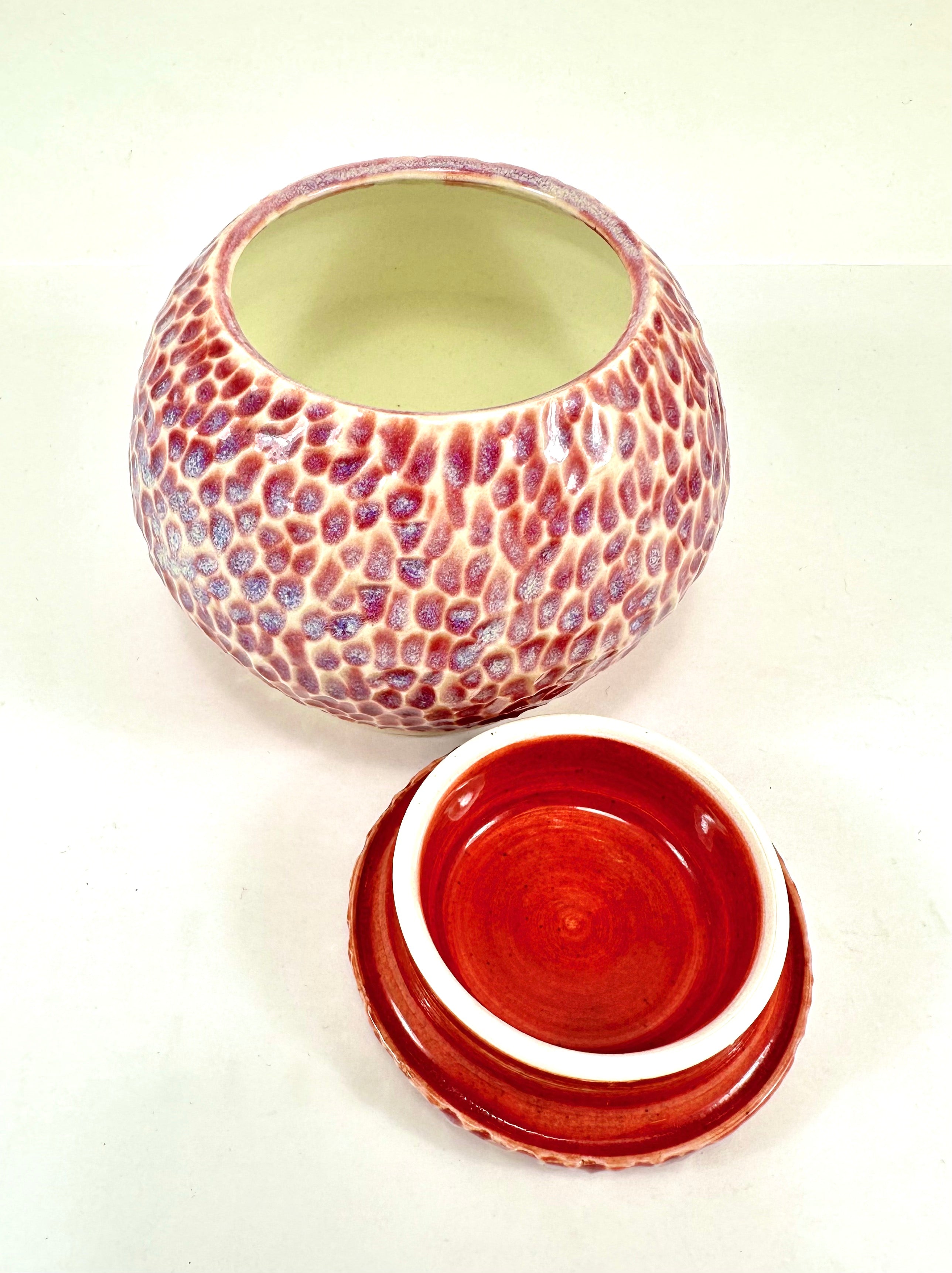 A handmade ceramic sugar jar, The sugar jar is glazed in a beautiful pink, blue, beige and red glaze. It is carved all over to add texture. The sugar jar is round with a flat lid.