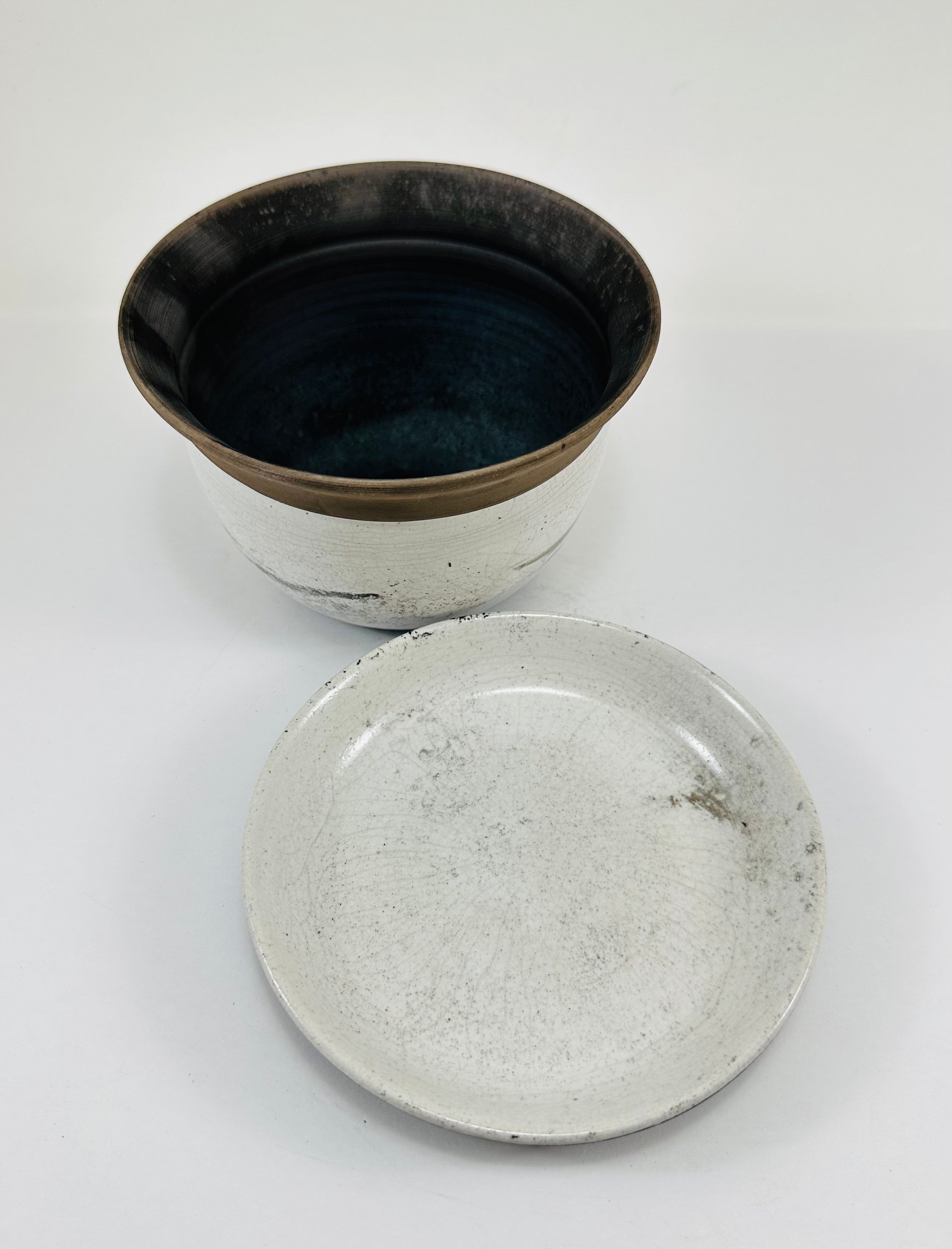A hand made ceramic planter and plate with a stunning, matte Raku glaze. The colours show whites and blacks. The planter is bowl shaped and has a flared opening. The top rim of the planter is black and the bottom body and plate is white.