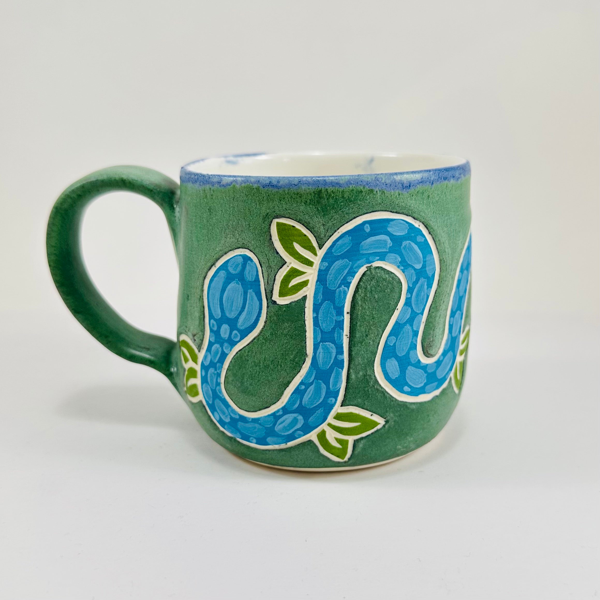 A handmade ceramic mug with a blue snake illustration on it. The mug is glazed in a beautiful robins egg green on the outside and white on the inside. The mug is made from porcelain and has a smooth comfortable handle.