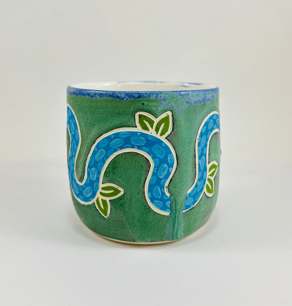 A handmade ceramic mug with a blue snake illustration on it. The mug is glazed in a beautiful robins egg green on the outside and white on the inside. The mug is made from porcelain and has a smooth comfortable handle.