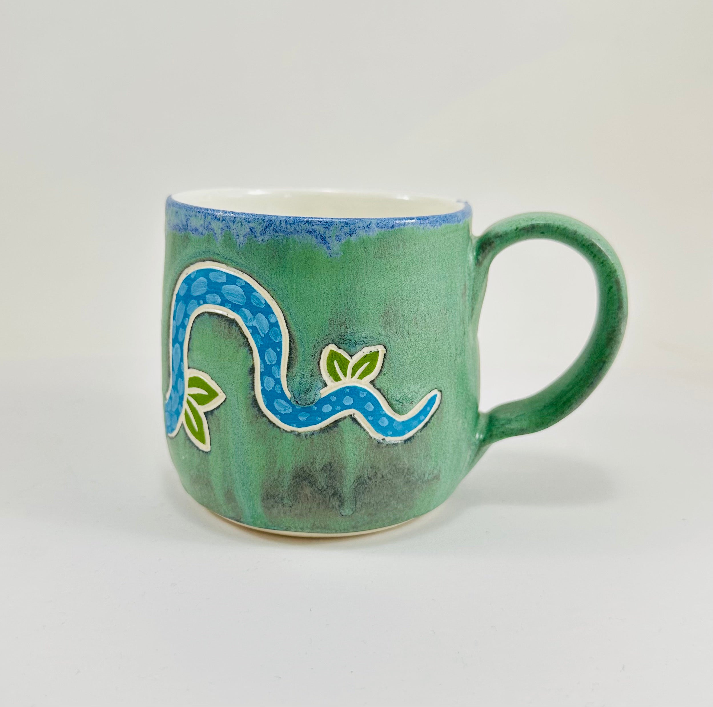 A handmade ceramic mug with a blue snake illustration on it. The mug is glazed in a beautiful robins egg green on the outside and white on the inside. The mug is made from porcelain and has a smooth comfortable handle.