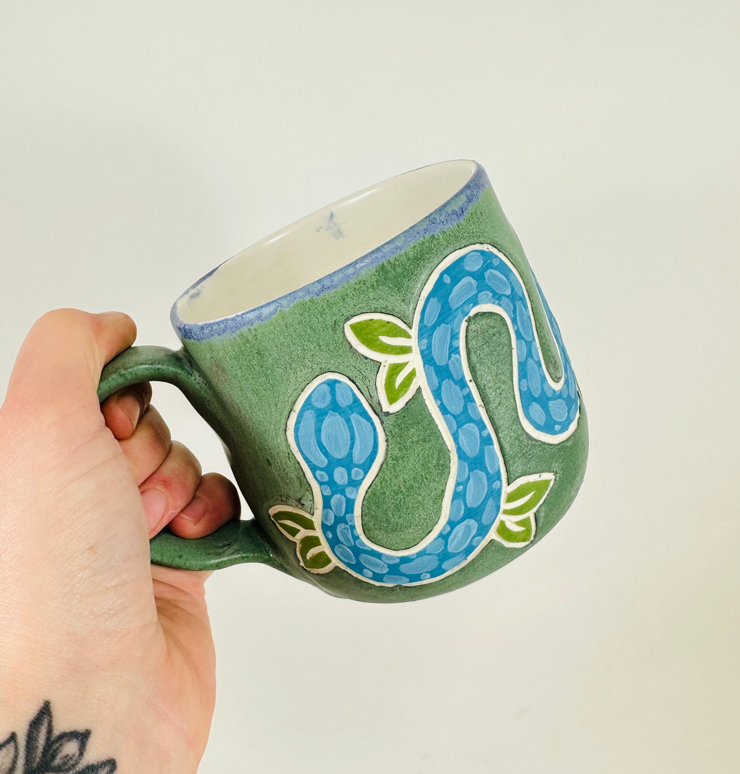 A handmade ceramic mug with a blue snake illustration on it. The mug is glazed in a beautiful robins egg green on the outside and white on the inside. The mug is made from porcelain and has a smooth comfortable handle.
