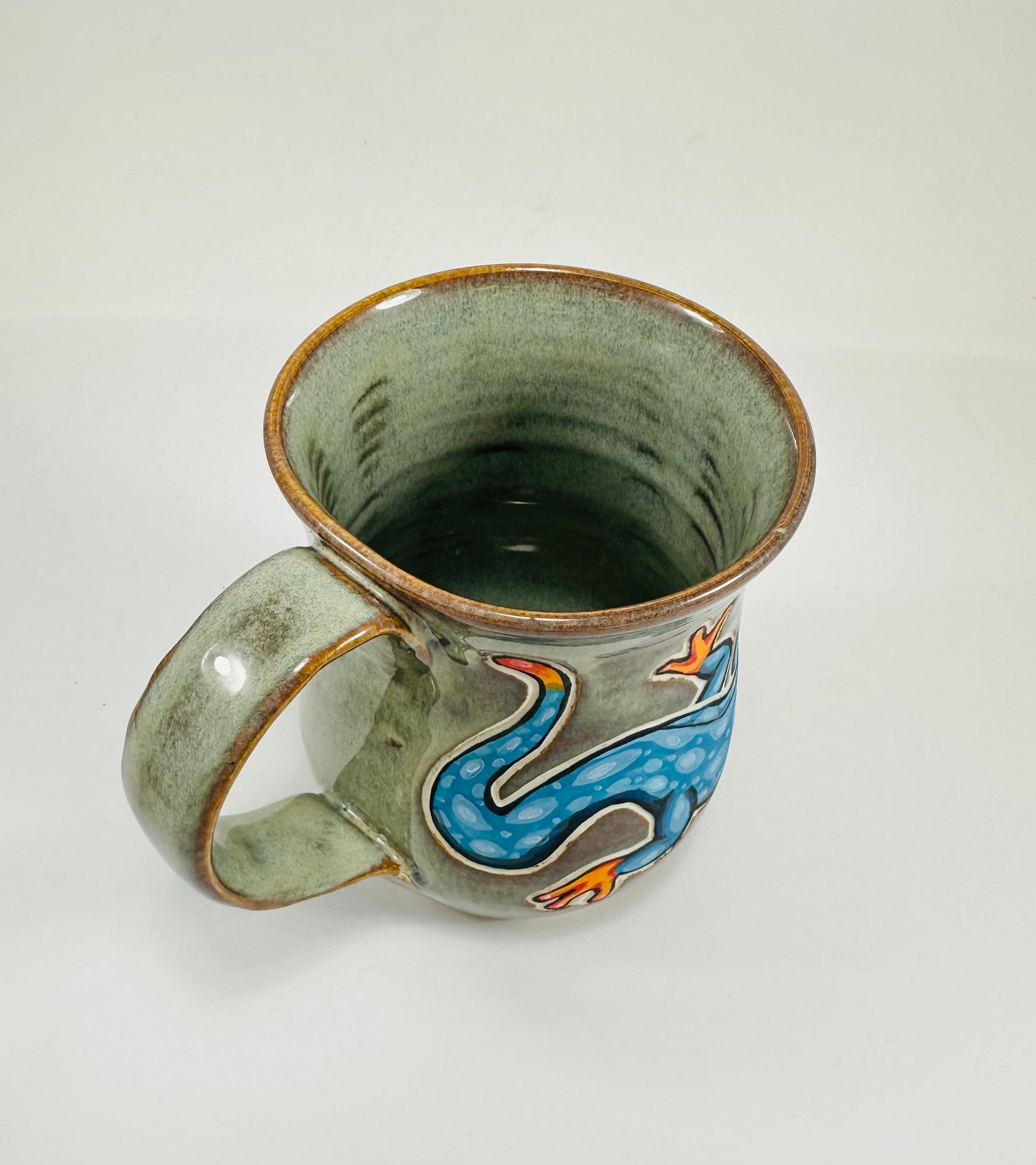 A handmade ceramic mug with a blue lizard illustration on it. The mug is made from porcelain clay and is glazed in a beautiful green glaze.