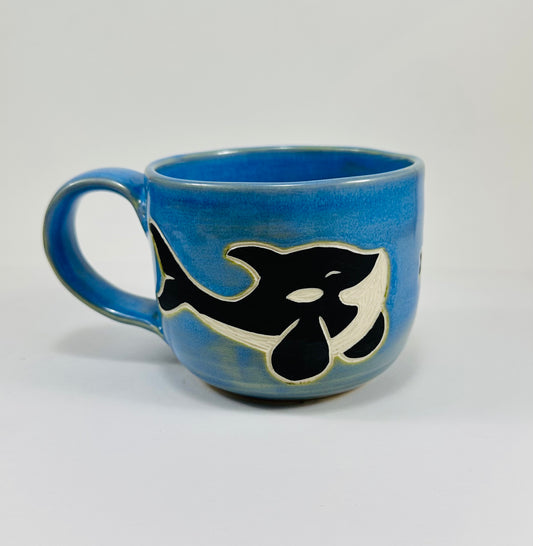A handmade ceramic mug with an orca chasing fish illustration on it. The mug is made from porcelain clay and has a beautiful blue glaze.