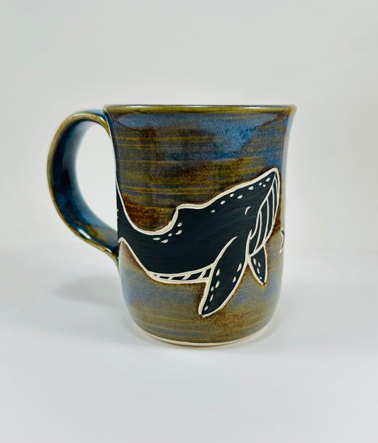 A handmade ceramic mug with a blue whale illustration on it and fish. The mug is made from porcelain clay and has a beautiful blue and brown glaze.
