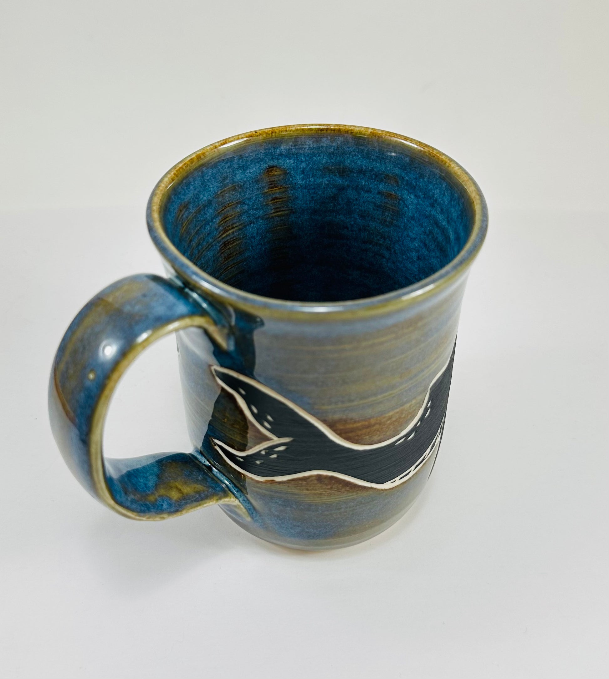 A handmade ceramic mug with a blue whale illustration on it and fish. The mug is made from porcelain clay and has a beautiful blue and brown glaze.