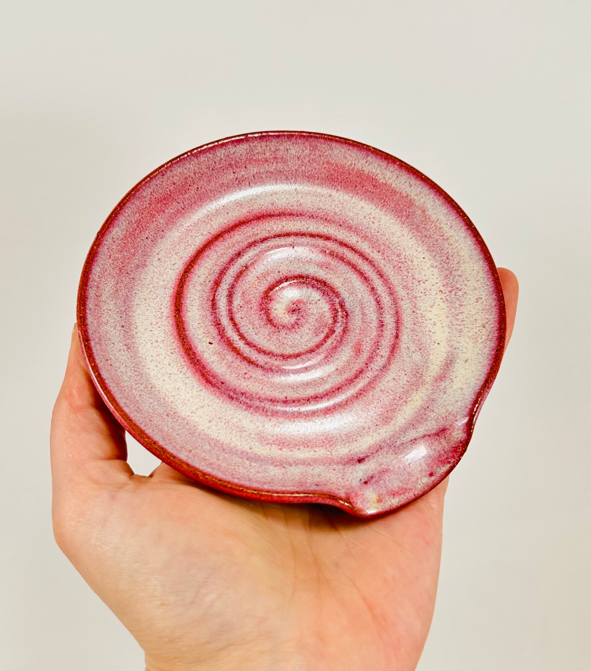 A handmade ceramic spoon rest in a beautiful pink glaze on red clay.