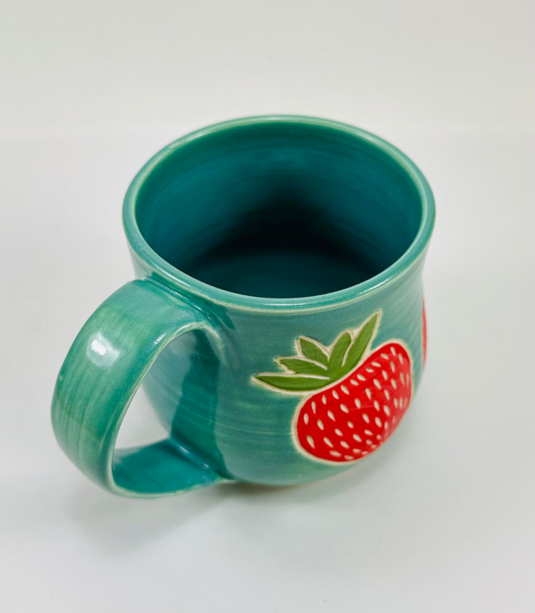 handmade ceramic mug in a beautiful blue/green glaze and illustrations of strawberries on it