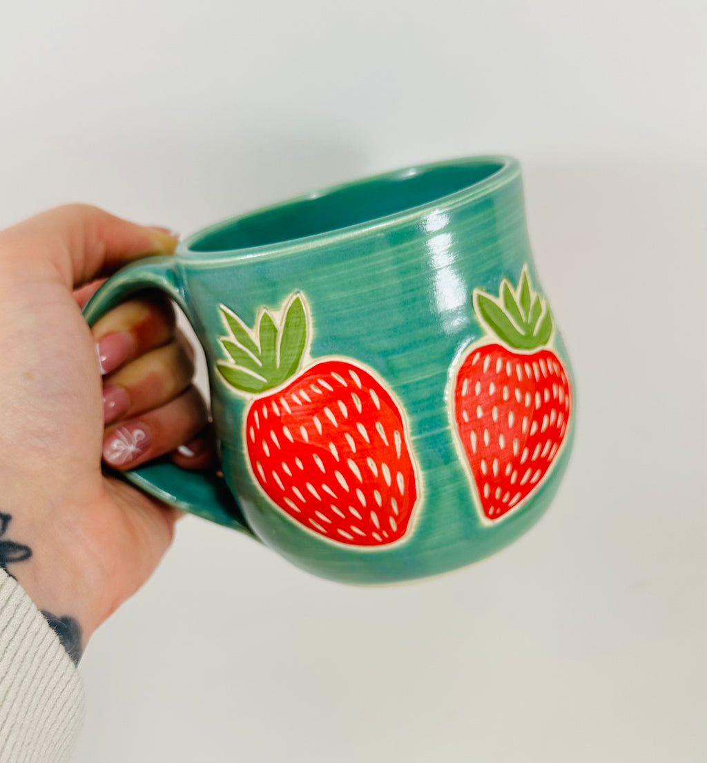 handmade ceramic mug in a beautiful blue/green glaze and illustrations of strawberries on it
